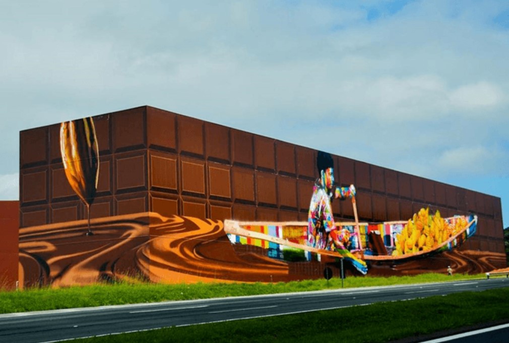 Fábrica da Cacau Show, que tem um mural lindo feito pelo artista Kobra, ilustrando um homem dentro de uma canoa remando em um lado de chocolate. Todo colorido.